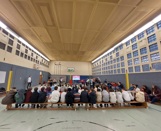 KI generiert: Das Bild zeigt eine Gruppe von Menschen, die auf Bänken in einer Turnhalle sitzen, während mehrere Personen vorne an einer Bühne oder einem Präsentationsbereich stehen. Im Hintergrund ist ein Bildschirm mit einer Präsentation zu sehen.