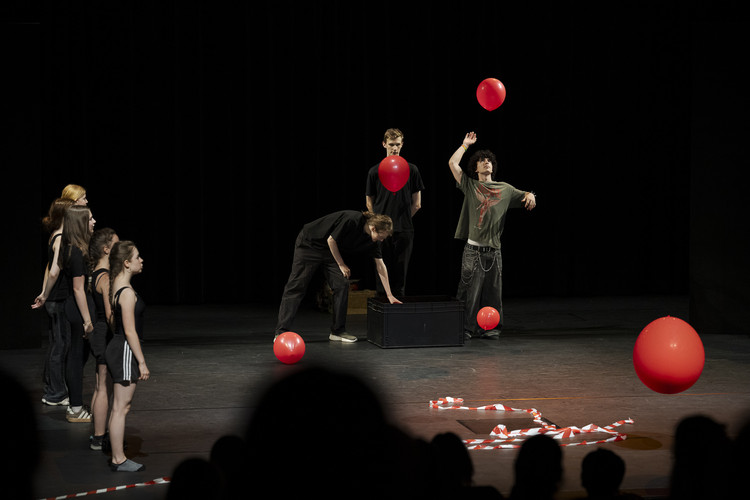 KI generiert: Das Bild zeigt eine Gruppe von Personen auf einer Bühne, die mit roten Luftballons und einem schwarzen Kasten interagieren. Einige Personen schauen zu, während andere mit den Requisiten agieren.