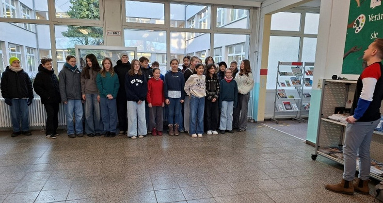 KI generiert: Eine Gruppe von Personen steht in einem Innenbereich, möglicherweise in einer Schule, und eine Person vorne rechts scheint etwas zu präsentieren oder zu erklären. Im Hintergrund sind große Fenster und ein Bücherregal sichtbar.