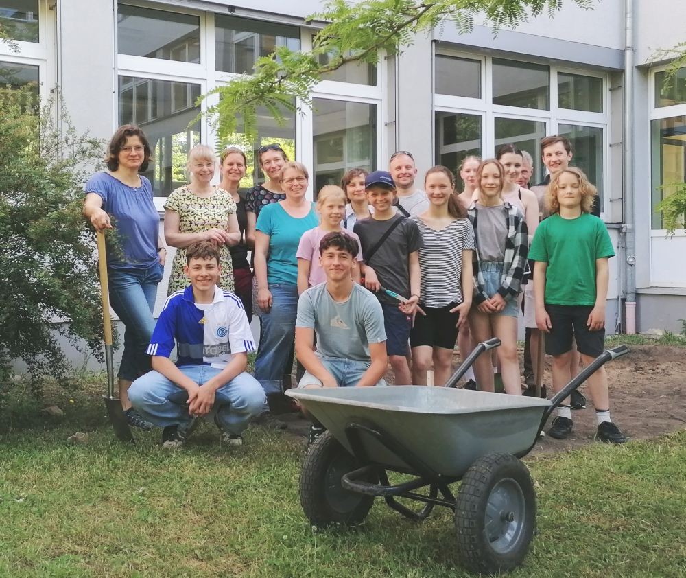 KI generiert: Das Bild zeigt eine Gruppe von Menschen, die sich mit Gartenwerkzeugen in einem Außenbereich vor einem Gebäude versammelt haben. Im Vordergrund steht eine Schubkarre, was auf ein gemeinschaftliches Garten- oder Bauprojekt hindeutet.