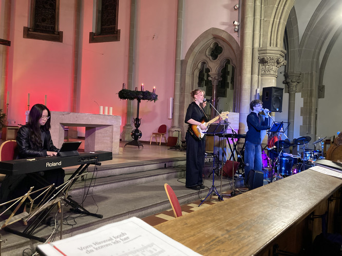 KI generiert: Das Bild zeigt eine musikalische Darbietung in einer Kirche, bei der eine Person ein Keyboard spielt und zwei Personen singen bzw. ein Instrument wie eine Gitarre spielen. Im Hintergrund sind ein Altar und ein Adventskranz sichtbar.