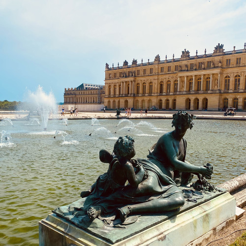 KI generiert: Das Bild zeigt eine Bronzeskulptur vor dem prächtigen Schloss Versailles, das am Rande eines Sees mit sprudelnden Springbrunnen liegt. Im Hintergrund sind die opulente Architektur und einige vorbeigehende Menschen zu sehen.