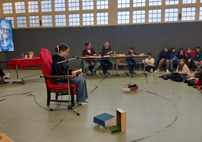 KI generiert: Ein Mädchen liest aus einem Buch auf einer Bühne, während um sie herum eine Jury und ein Publikum sitzen. Der Raum wirkt wie eine Halle, und im Hintergrund sind Bücher und ein großes Bild sichtbar.