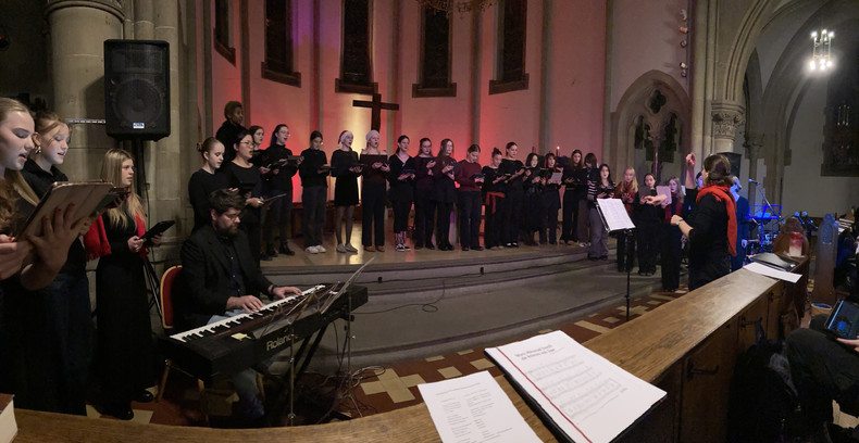 KI generiert: Das Bild zeigt einen Chor, der in einer Kirche oder einem sakralen Raum auftritt, begleitet von einem Pianisten und unter der Leitung eines Dirigenten. Die Szenerie ist stimmungsvoll beleuchtet, und im Hintergrund ist ein Kreuz zu sehen.