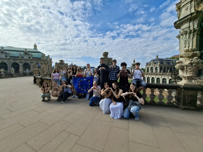 KI generiert: Eine Gruppe von Menschen posiert auf einem historischen Platz vor einer beeindruckenden Architektur und hält eine EU-Flagge. Der Himmel ist blau mit leichten Wolken.