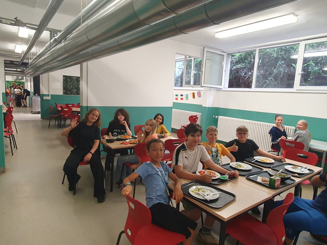 KI generiert: Das Bild zeigt eine Gruppe von Kindern, die in einer Kantine an mehreren Tischen sitzen und essen. Die Atmosphäre wirkt fröhlich und gesellig.