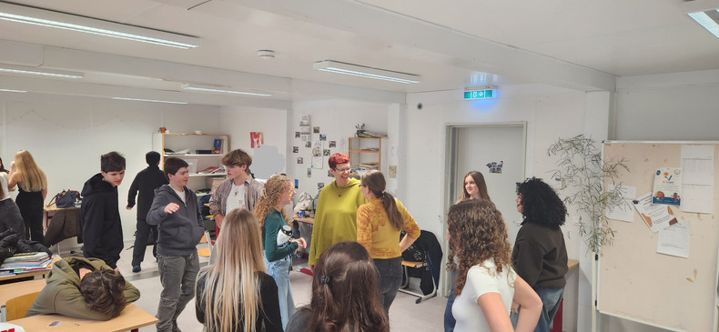 KI generiert: Das Bild zeigt eine Gruppe von Menschen in einem Klassenraum, die miteinander sprechen. Im Vordergrund sind mehrere Personen in einem Kreis angeordnet, während im Hintergrund andere Personen ebenfalls interagieren.