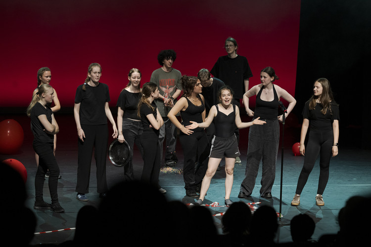 KI generiert: Eine Gruppe von Menschen in schwarzer Kleidung steht auf einer Bühne mit einem roten Hintergrund. Sie scheinen in einer Theater- oder Performance-Situation zu sein.