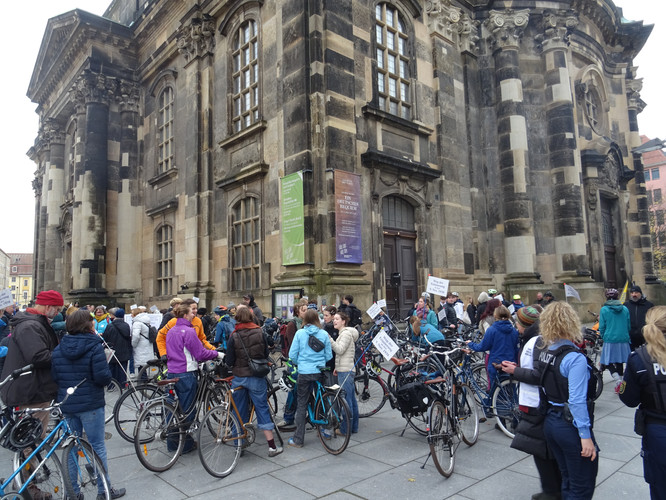 KI generiert: Das Bild zeigt eine Gruppe von Menschen mit Fahrrädern vor einem historischen Gebäude, die an einer Versammlung oder Protest teilnehmen. Einige Teilnehmer halten Schilder, und es sind Polizeibeamte anwesend.