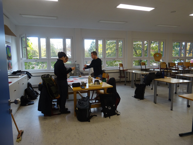 KI generiert: Das Bild zeigt einen Raum mit zwei Personen, die an einem Tisch stehen und offenbar miteinander interagieren. Der Raum hat viele Fenster, durch die viel Tageslicht hereinkommt, und es sind mehrere Tische und Stühle zu sehen.