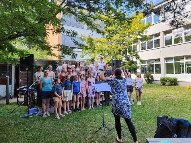 KI generiert: Ein Chor bestehend aus Kindern und Jugendlichen singt unter freiem Himmel, während eine Dirigentin sie leitet. Im Hintergrund ist eine Schulgebäude und grüne Bäume zu sehen.