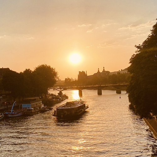 KI generiert: Das Bild zeigt einen Fluss bei Sonnenuntergang, über den eine Brücke führt, mit einem Schiff im Vordergrund. Die Szenerie ist von Bäumen gesäumt und strahlt eine warme, friedliche Atmosphäre aus.