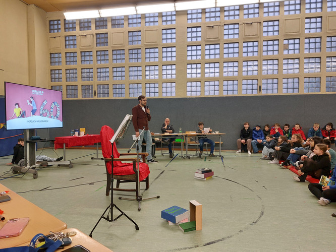 KI generiert: Eine Person hält in einer Turnhalle eine Präsentation oder Vorlesung vor einer Gruppe sitzender Zuschauer. Ein Bildschirm zeigt den Text "Herzlich Willkommen".