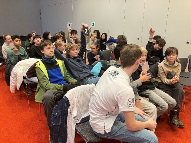 KI generiert: Das Bild zeigt eine Gruppe von Schülern, die in einem Raum auf Stühlen sitzen, einige heben nummerierte Schilder in die Luft. Die meisten Jugendlichen schauen aufmerksam in die gleiche Richtung, was auf eine gemeinsame Aktivität oder Präsentation hindeutet.