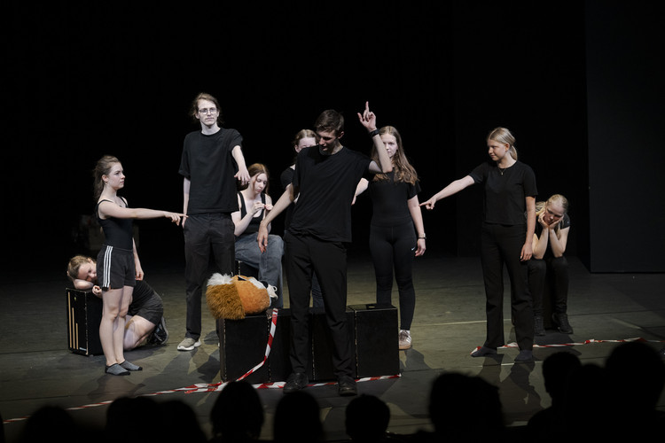KI generiert: Das Bild zeigt eine Gruppe von Menschen auf einer Theaterbühne, die in schwarzer Kleidung stehen und performen, während sie unterschiedliche emotionale Posen einnehmen. Einige von ihnen zeigen mit Fingern auf jemanden in der Mitte, was auf eine dramatische Szene hindeutet.