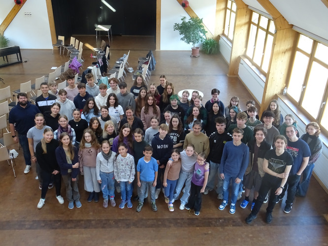KI generiert: Eine große Gruppe von Menschen, hauptsächlich Jugendliche, steht zusammen in einem Raum mit Stuhlreihen und großen Fenstern. Sie blicken alle zur Kamera und wirken fröhlich.