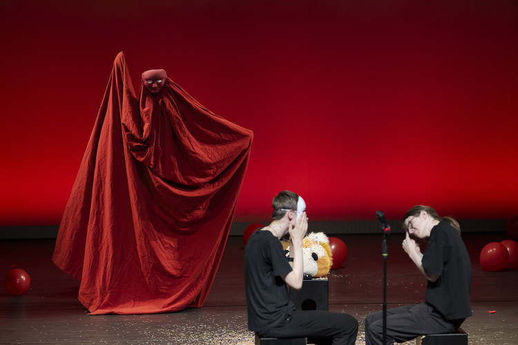 KI generiert: Das Bild zeigt eine künstlerische Performance auf einer Bühne. Ein Darsteller in einem langen, roten Kostüm mit Maske steht, während zwei weitere Personen maskiert auf dem Boden sitzen, umgeben von roten Luftballons und Requisiten.