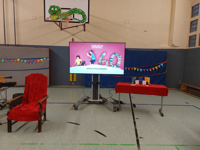 KI generiert: Das Bild zeigt einen Raum, der offenbar für einen Vorlesewettbewerb vorbereitet ist, inklusive eines Bildschirms mit der Aufschrift "Vorlesewettbewerb", einem dekorierten Tisch und einem roten Stuhl. Im Hintergrund ist eine Basketballkorbanlage mit einer darauf gemalten Raupe sichtbar.