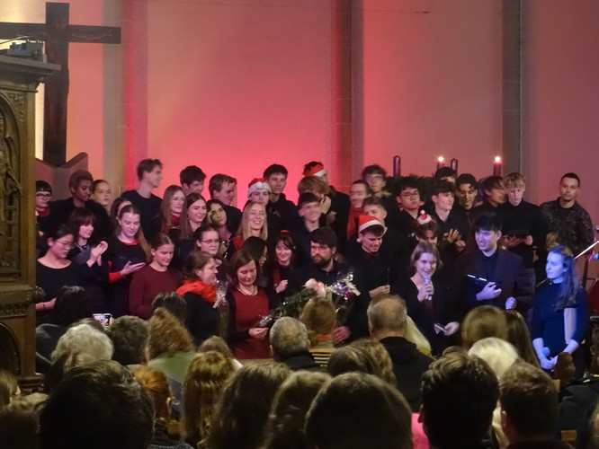 KI generiert: Das Bild zeigt eine Gruppe von Menschen, die in einer Kirche vor einem Publikum stehen, einige halten Blumen in den Händen. Die Aufnahme vermittelt eine festliche Stimmung, möglicherweise während einer Feier oder eines Konzerts.