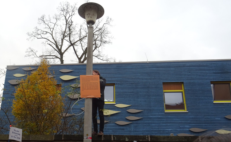KI generiert: Ein Mensch steht auf einer Mauer und hängt ein Schild an eine Straßenlaterne vor einem blauen Gebäude. Im Hintergrund sind herbstliche Bäume und ein kunstvolles Fassadenmuster zu sehen.