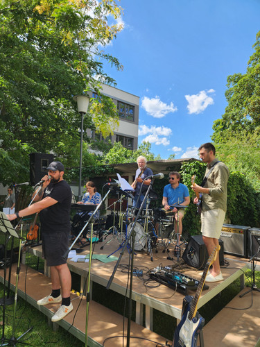 KI generiert: Eine Band spielt auf einer kleinen Freiluftbühne, umgeben von grünem Laub und einem sonnigen Himmel. Die Bandmitglieder sind mit verschiedenen Instrumenten aktiv, darunter eine Gitarre, ein Schlagzeug und ein Keyboard.