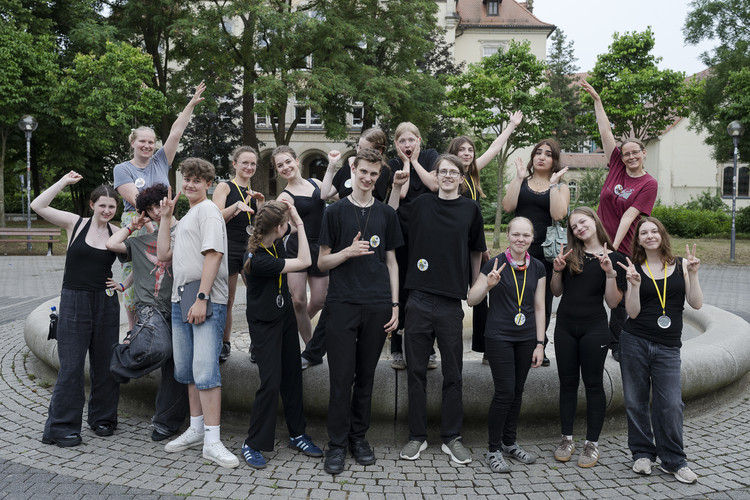 KI generiert: Eine Gruppe von Menschen posiert fröhlich in einem öffentlichen Platz auf einem großen Brunnen. Sie tragen Medaillen und scheinen eine feierliche oder glückliche Veranstaltung zu feiern.