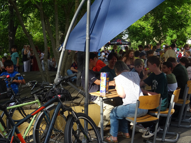 KI generiert: Das Bild zeigt eine lebhafte Outdoor-Veranstaltung, bei der Menschen an Tischen sitzen und sich unterhalten, umgeben von Fahrrädern und Bäumen. Im Hintergrund sind weitere Teilnehmer und Bäume zu sehen, was auf ein geselliges, sommerliches Treffen im Freien hindeutet.