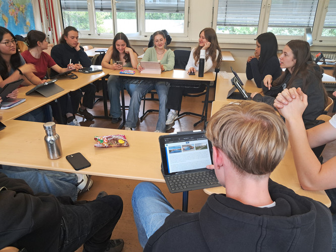 KI generiert: Eine Gruppe von Schülern sitzt in einem Klassenraum in einer U-Form zusammen, wobei einige Tablets verwenden. Sie scheinen an einer Diskussion oder Gruppenarbeit teilzunehmen.