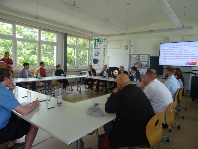 KI generiert: Das Bild zeigt eine Gruppe von Menschen, die in einem Konferenzraum um einen Tisch versammelt sind, möglicherweise um ein Meeting oder eine Diskussion abzuhalten. Auf einem Bildschirm an der Wand werden Informationen über ein Bildungsprogramm präsentiert.